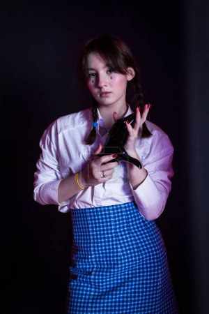Photo for A cute girl with drawn tears, dressed in a white shirt and blue plaid skirt, holds black plastic hand in a dark room. - Royalty Free Image