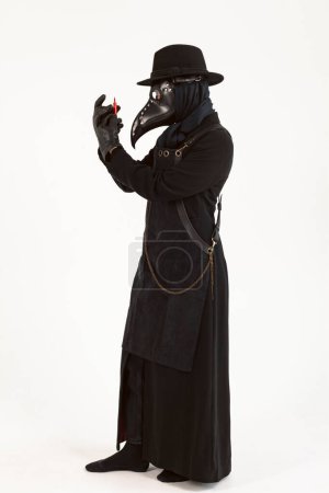 Full-length portrait of a Plague doctor holding a syringe and needle with Medicine or serum, antidote. high-quality costume. Isolated on a white background. COVID-19, epidemic and pandemic concept.