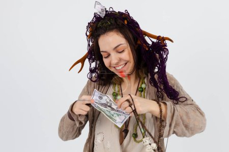 Photo for A portrait of a female druid or shaman with horns, holding ritual talismans and money cash in her hands on a white isolated background. Shamanic practices, Spiritualism and materialism. - Royalty Free Image