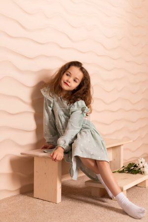 Photo for Portrait of a charming little girl, well dressed in a pastel green dress with a slight makeup and brown loose hair posing with a flowers on a short bench against a wavy beige wall. - Royalty Free Image