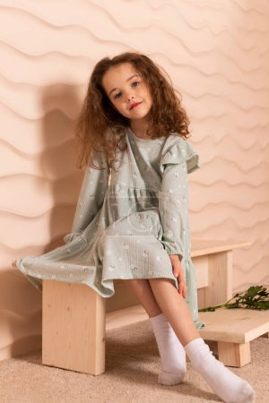Photo for Portrait of a charming little girl, well dressed in a pastel green dress with a slight makeup and brown loose hair posing with a flowers on a short bench against a wavy beige wall. - Royalty Free Image