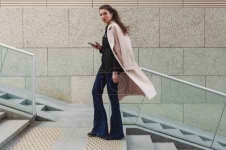 Photo for A beautiful businesswoman with a white coat draped over her shoulders climbs steps on a street in the business district with a phone in her hand. - Royalty Free Image
