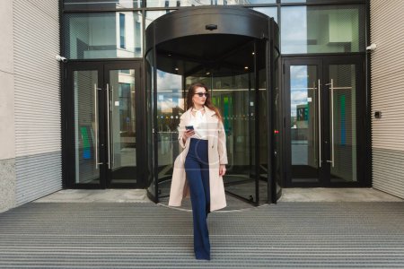 Photo for A beautiful businesswoman in a light beige coat and black sunglasses exits a glazed building in the business district with a phone in her hands. - Royalty Free Image