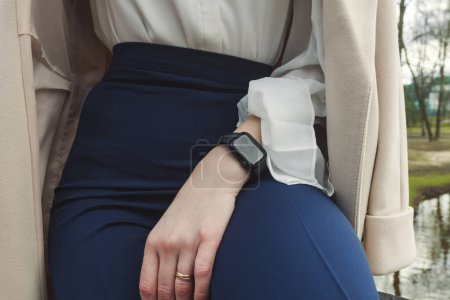 Photo for Fashionable unrecognizable girl sitting on the rails of a bridge over a creek in the park, with a coat thrown over her shoulders, smart watch on her wrist, stylish tight dark blue pants, white shirt. - Royalty Free Image