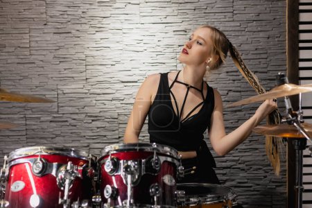 Photo for Young cute blonde girl in wireless earphones sitting by the drums in a garage. female drummer. musician. - Royalty Free Image