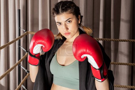 Photo for A beautiful confident girl in red boxing gloves stands in a fighting stance and looks into the camera in a boxing ring. - Royalty Free Image