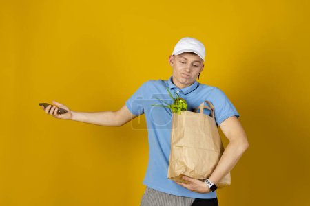 Téléchargez les photos : Un jeune homme portant un t-shirt et une casquette bleus tient un sac en papier avec de la nourriture. Isolé sur fond jaune. Concept de livraison. - en image libre de droit