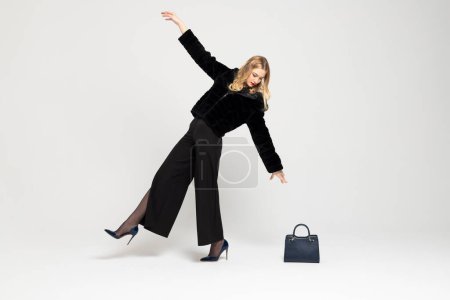 Photo for Young Curly blonde girl elegantly dressed, leaning back and reaching her hand to a handbag on the floor. expensive clothes, style and fashion concept. - Royalty Free Image