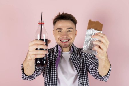 Foto de Un joven feliz bebiendo coca y comiendo una barra de chocolate. comida rápida y adicción al azúcar, dieta poco saludable. Aislado sobre fondo rosa. - Imagen libre de derechos