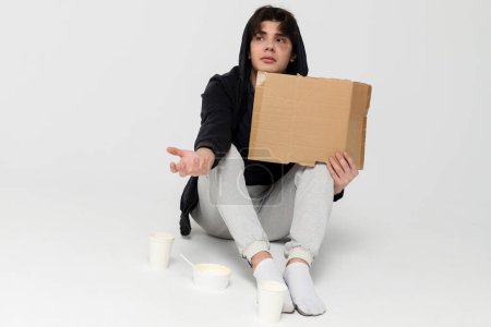 Photo for A homeless young person with bruises under their eyes in dirty, tattered clothing, holding out a hand and empty blank cardboard sign. Isolated on white background. - Royalty Free Image