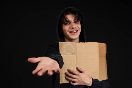 Photo for A smiling homeless young person with bruises under their eyes in dirty, tattered clothing, holding out an empty blank cardboard sign and hand. Isolated on black background. - Royalty Free Image