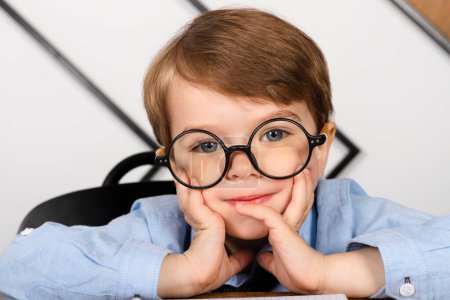 Photo for Little boy in round eyeglasses, blue shirt and tie sitting at the desk in the office with a notepad. Smart little kid, Child prodigy. - Royalty Free Image