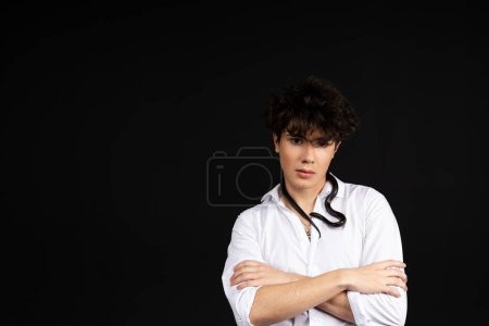 Foto de Joven hombre guapo con una camisa blanca sentado en una silla con una serpiente negra arrastrándose alrededor de su cuello. Aislado sobre fondo negro. - Imagen libre de derechos