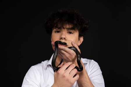 Foto de Joven hombre guapo en camisa blanca con una serpiente negra arrastrándose en su brazo o mano. Aislado sobre fondo negro. - Imagen libre de derechos