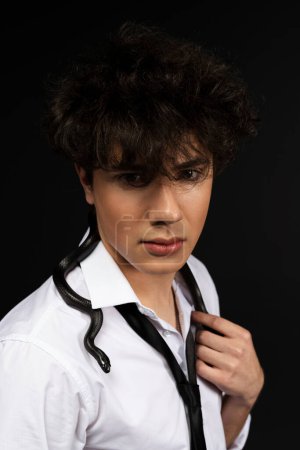 Foto de Joven hombre guapo en camisa blanca con una serpiente negra arrastrándose sobre su hombro. Aislado sobre fondo negro. - Imagen libre de derechos