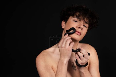 Foto de Joven hombre guapo con el torso desnudo, con una serpiente negra arrastrándose en sus brazos o manos. Aislado sobre fondo negro. - Imagen libre de derechos