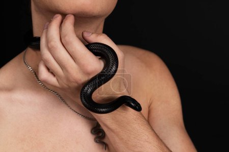 Photo for Young handsome man with naked torso, with a black snake crawling around his neck. Isolated on black background. - Royalty Free Image
