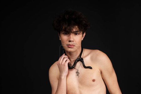 Photo for Young handsome man with naked torso, with a black snake crawling around his neck. Isolated on black background. - Royalty Free Image