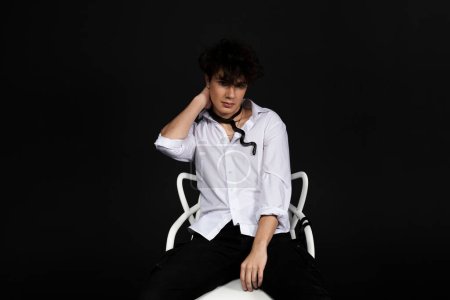Foto de Joven hombre guapo con una camisa blanca sentado en una silla con una serpiente negra arrastrándose alrededor de su cuello. Aislado sobre fondo negro. - Imagen libre de derechos