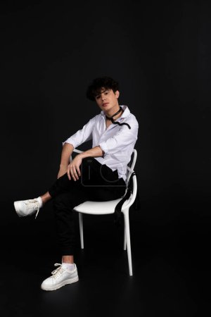 Photo for Young handsome man in a white shirt sitting on a chair with a black snake crawling around his neck. Isolated on black background. - Royalty Free Image