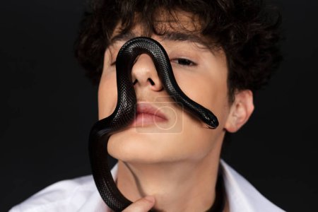 Foto de Joven hombre guapo con una camisa blanca con una serpiente negra gateando en su cara. Aislado sobre fondo negro. - Imagen libre de derechos