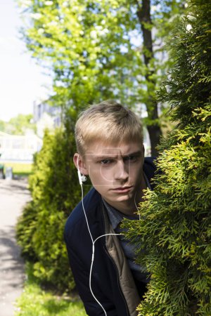 Photo for Young blond man in casual clothes and earphones peeking from a bush in the park. - Royalty Free Image