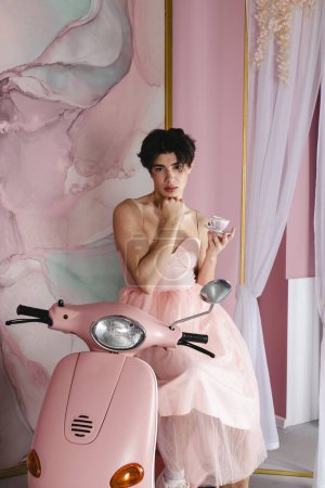 Foto de Retrato de un gay tierno en un vestido rosa sentado en una moto o ciclomotor con una taza de té. Glamour homosexual joven chico con maquillaje posando en un traje femenino. - Imagen libre de derechos