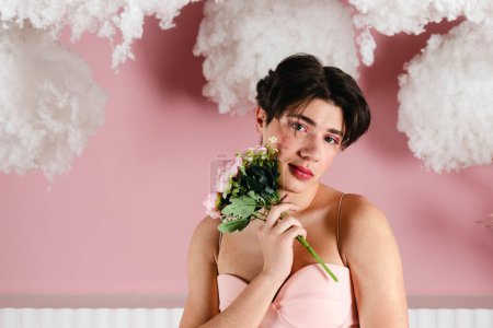 Foto de Close-up Retrato de un gay tierno en un vestido rosa con flores, ramo. Joven homosexual glamour con maquillaje posando en un traje femenino. disparo en la cabeza. - Imagen libre de derechos