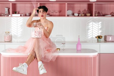 Foto de Retrato de un gay tierno en un vestido rosa en una cocina. Glamour homosexual joven chico con maquillaje posando en un traje femenino. - Imagen libre de derechos