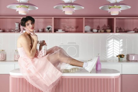 Foto de Retrato de un gay tierno en un vestido rosa en una cocina. Glamour homosexual joven chico con maquillaje posando en un traje femenino. - Imagen libre de derechos