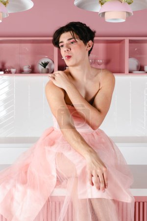 Foto de Retrato de un gay tierno en un vestido rosa con una piruleta. Glamour homosexual joven chico con maquillaje posando en un traje femenino. - Imagen libre de derechos