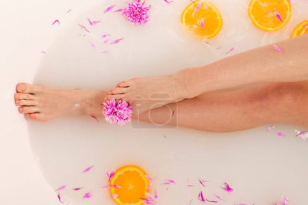 Téléchargez les photos : Jambes tendres de fille dans un bain rempli de tranches de fruits orange et de pétales de fleurs. Concept beauté et soin de la peau. Vue du dessus. - en image libre de droit