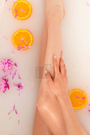 Téléchargez les photos : Jambes tendres de fille dans un bain rempli de tranches de fruits orange et de pétales de fleurs. Concept beauté et soin de la peau. Vue du dessus. - en image libre de droit
