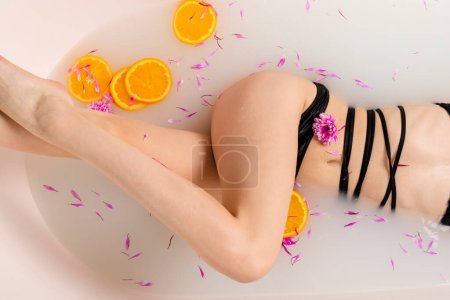 Téléchargez les photos : Corps de fille tendre dans un bain à moitié rempli d'eau avec des tranches d'orange de fruits et des pétales de fleurs. Concept beauté et soin de la peau. Vue du dessus. - en image libre de droit