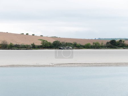 Photo for Sandy seashore on a cloudy day. Calm water surface. The remains of an old ship on the shore. A plowed field. Minimalistic landscape. - Royalty Free Image