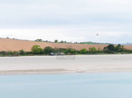 Photo for Seaside landscape and a farmer's field on a hill. Sandy seashore on a cloudy day. Calm water surface. The remains of an old ship on the shore. A plowed field. - Royalty Free Image