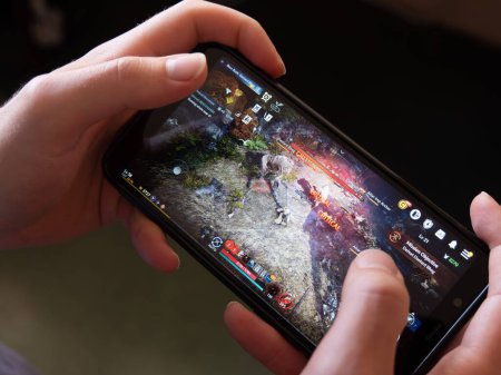 Téléchargez les photos : Cork, Ireland, July 31, 2022. A mobile phone in the hands of a white man. Playing mobile MMORPG Black Desert Mobile. A Korean mobile game on a smartphone screen, illustrative editorial. - en image libre de droit