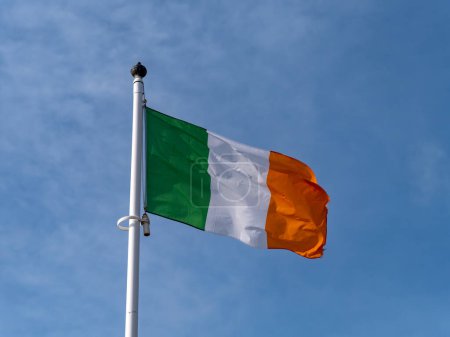Foto de Una bandera de Irlanda en un asta de la bandera contra un cielo azul claro. - Imagen libre de derechos