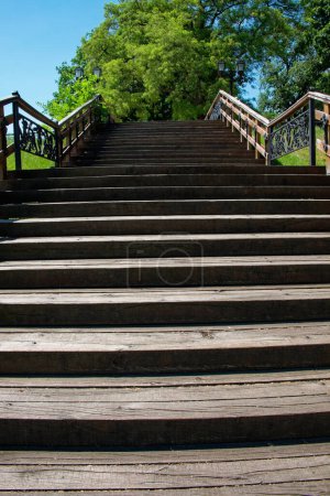 Foto de Esta es una foto de una escalera de madera en un parque con una barandilla de metal, rodeado de árboles bajo un cielo azul. - Imagen libre de derechos