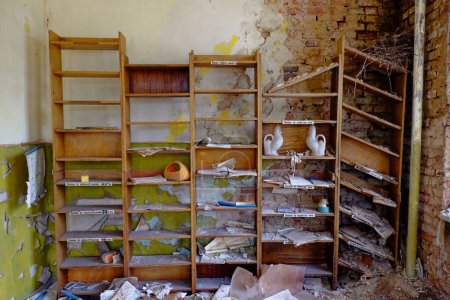 Téléchargez les photos : Une pièce abandonnée avec une étagère en bois remplie de vieux livres et d'objets, et un mur de briques en ruine. Vieilles étagères pour les papiers dans une pièce abandonnée. Déchets et papiers éparpillés dans la pièce. - en image libre de droit