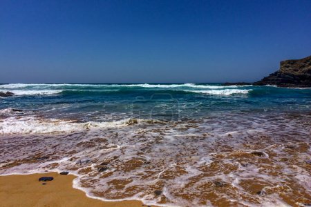 Foto de Una playa de arena con olas espumosas, y acantilados rocosos bajo un cielo azul claro. - Imagen libre de derechos