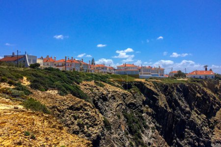 Coastal town with orange-roofed houses on a cliff, rocky terrain in the foreground, and a clear blue sky.