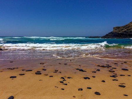 Foto de Las olas chocan en una playa soleada con acantilados en el fondo; arena. - Imagen libre de derechos