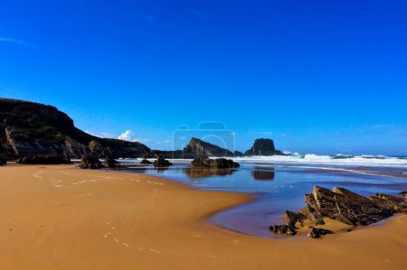 Una playa de arena con afloramientos rocosos, olas que se estrellan suavemente, bajo un cielo azul claro.