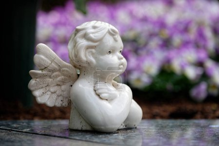 Téléchargez les photos : Une petite putto blanche sale sur une pierre tombale devant des fleurs printanières colorées en arrière-plan flou - en image libre de droit