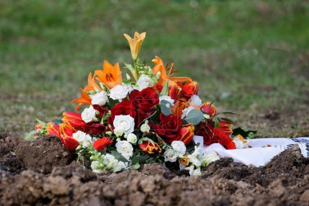 ein Blumengesteck mit weißen und roten Rosen, Feuerlilien und einem Trauerband liegt auf der Erde eines frischen Grabes auf einem Friedhof