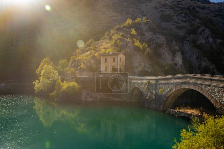 Der San Domenico-See in den Sagittario-Schluchten in den Abruzzen, L 'Aquila, Italien. Die kleine Einsiedelei mit der Steinbrücke. Die türkise Farbe des Wassers. Das Leuchten der Sonne, Fackeln bei Sonnenuntergang.