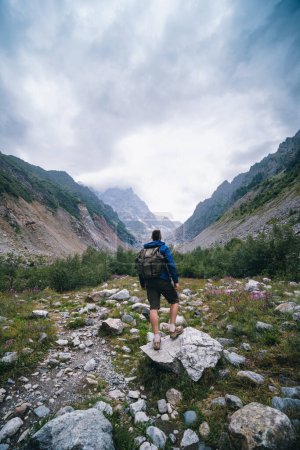 Téléchargez les photos : Voyageur homme avec un sac à dos debout sur une pierre, Sur le chemin, marcher sur la piste. Une vallée au milieu de hautes montagnes majestueuses.. Vue de dos. Le glacier Chalaadi, Géorgie, Caucase. Photo verticale. - en image libre de droit