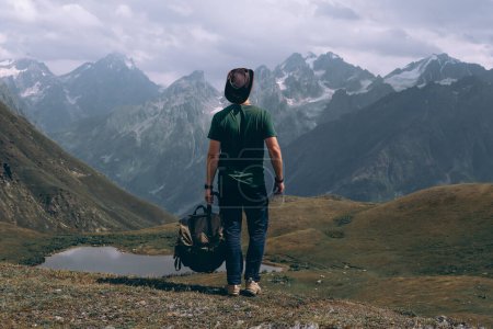 Téléchargez les photos : Un voyageur homme avec sac à dos et chapeau en cuir près du lac Koruldi, Mestia dans la chaîne de montagnes du Grand Caucase Upper Svaneti, Géorgie. Faune, voyage et vacances. Jour d'été. Vue arrière - en image libre de droit