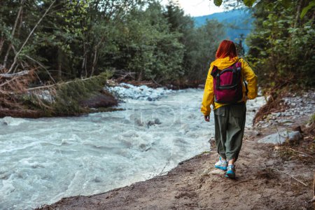 Foto de Una mujer excursionista en una chaqueta amarilla camina a lo largo de un sendero a lo largo de un río Dolra con un arroyo que corre, día de verano. Montañas concepto de viaje. Georgia - Imagen libre de derechos